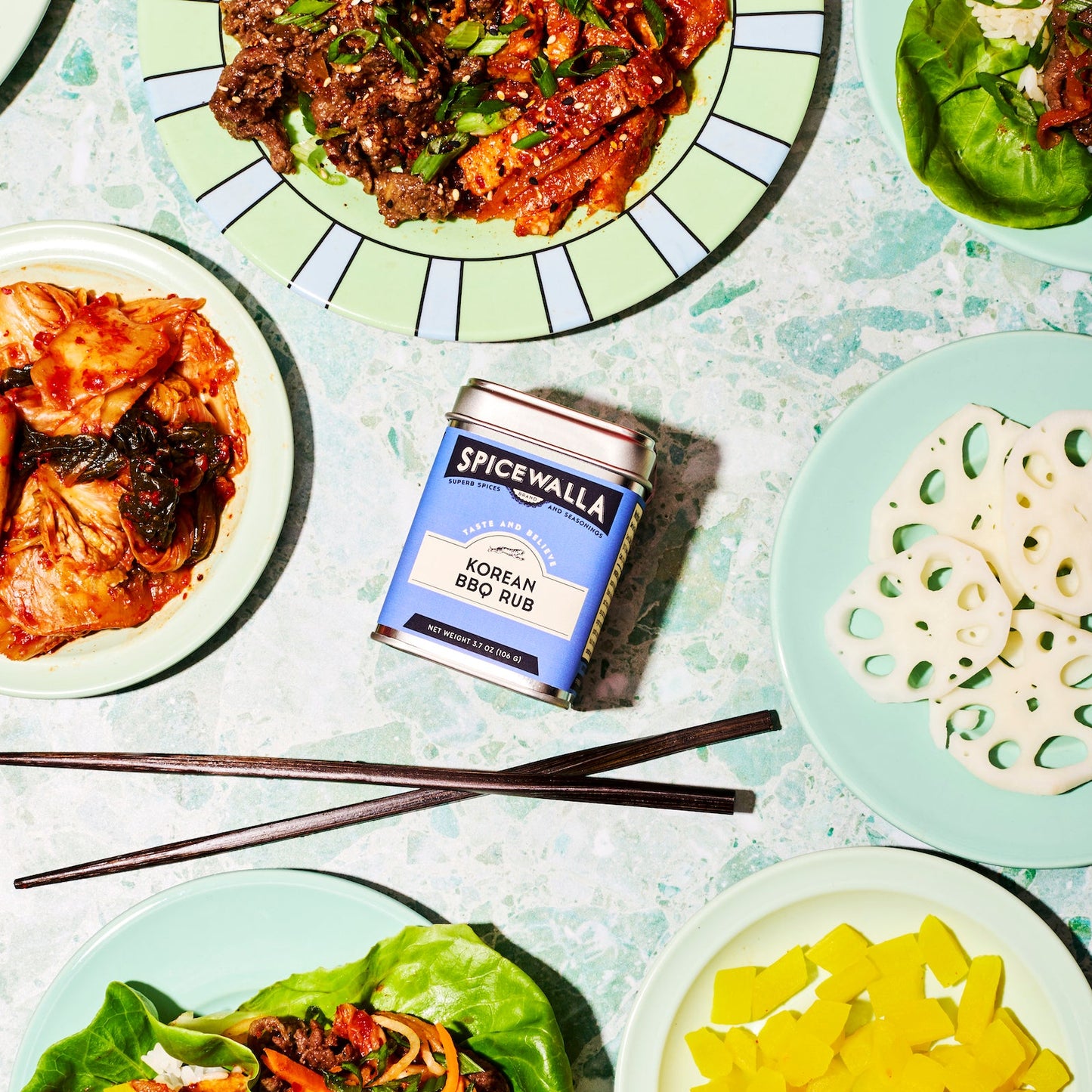 Spicewalla Korean BBQ Rub can with plates of food including kimchi and lettuce wraps on a light blue background.
