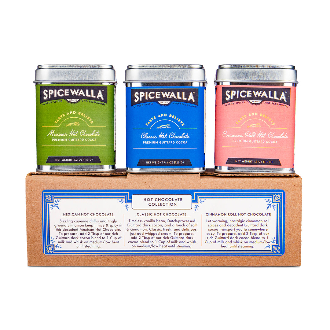 Three Spicewalla hot chocolate tins displayed on top of a cardboard gift box. The tins are labeled Mexican Hot Chocolate (green), Classic Hot Chocolate (blue), and Cinnamon Roll Hot Chocolate (pink).