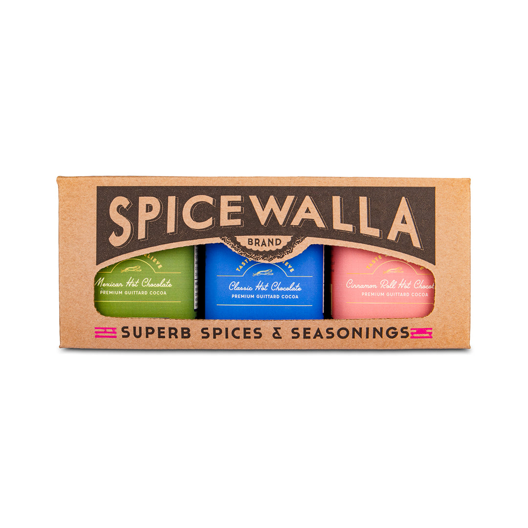 Spicewalla brand spice set in a brown cardboard box, containing three bottles of hot chocolate: green, blue, and pink, labeled 'Assam Hot Chocolate', 'Chilean Hot Chocolate', and 'Cinnamon Hot Chocolate'.