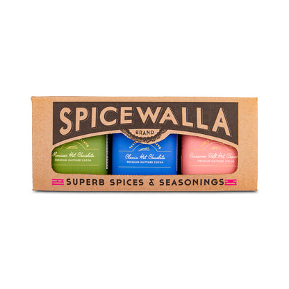 Spicewalla brand spice set in a brown cardboard box, containing three bottles of hot chocolate: green, blue, and pink, labeled 'Assam Hot Chocolate', 'Chilean Hot Chocolate', and 'Cinnamon Hot Chocolate'.