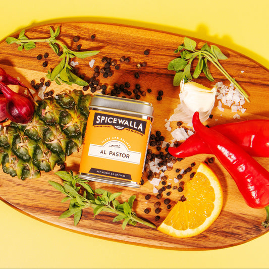 Spicewalla Al Pastor spice jar on a wooden cutting board with ingredients on a yellow background