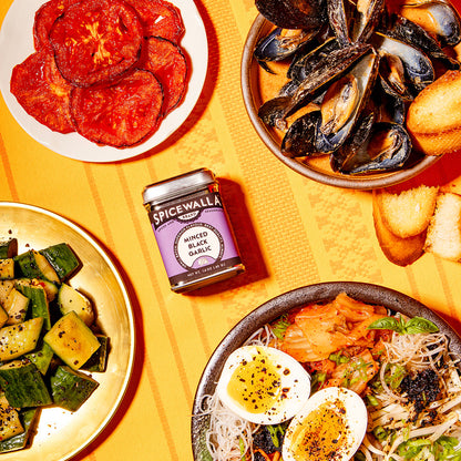 Assorted dishes including mussels, pickled vegetables, and a bowl of noodles on a yellow surface with a jar of Spicewalla sauce.