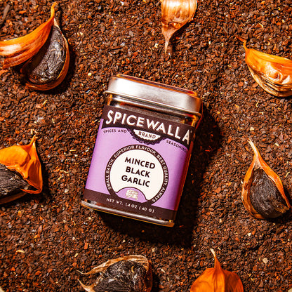Spicewalla minced black garlic jar on a bed of soil with garlic bulbs around it.