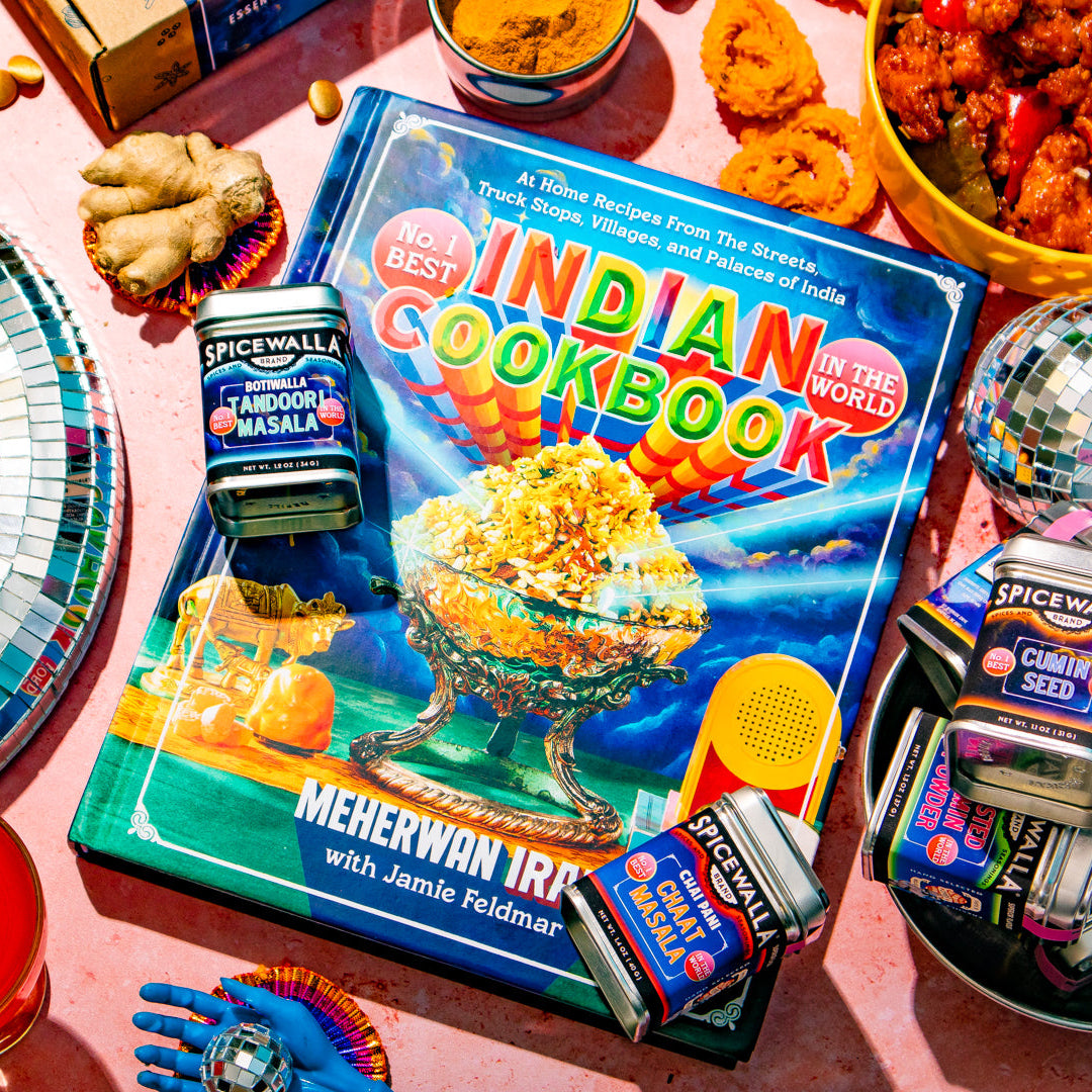 A colorful 'Indian Cookbook' book lies on a pink surface surrounded by food dishes, branded 'Spice Mall' tins, a disco ball, and other party props in a bright styled flat-lay scene.