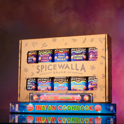 A SpiceWalla gift box filled with assorted spice jars, displayed on top of two Indian Cookbook volumes against a smoky purple and pink studio background.