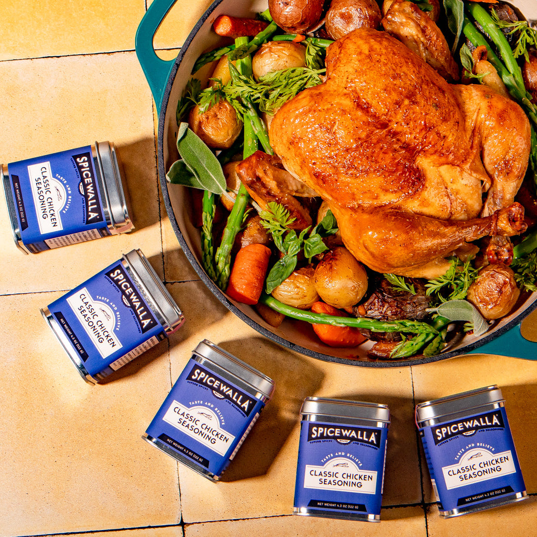 Roasted chicken with vegetables in a pan, surrounded by Spicewalla seasoning packets on a wooden surface.