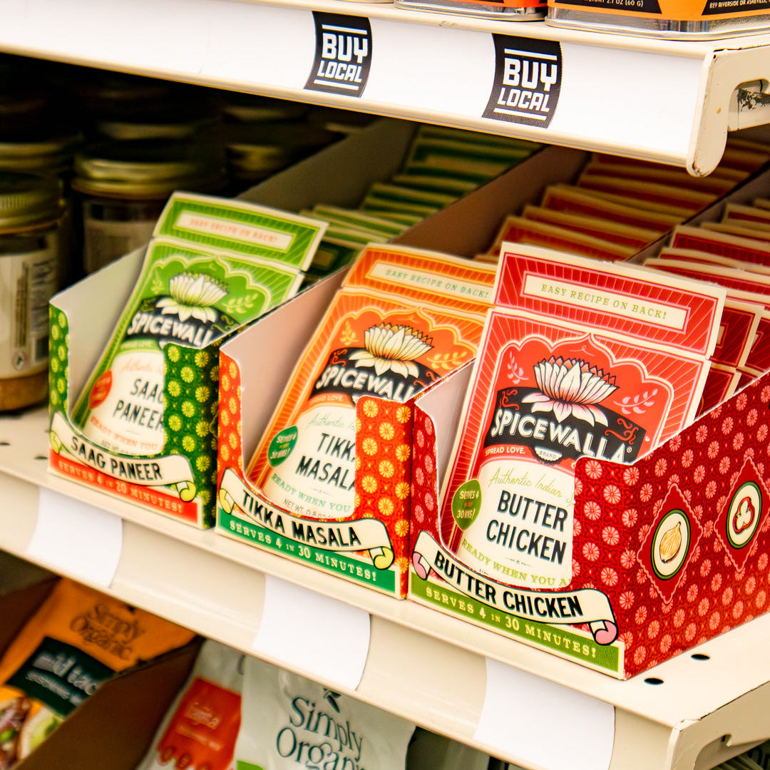 Spicewall seasoning and spice mix products displayed on store shelves, including Spicewalla seasoning tins on the top shelf and Saag Paneer, Tikka Masala, Butter Chicken pouches in front.