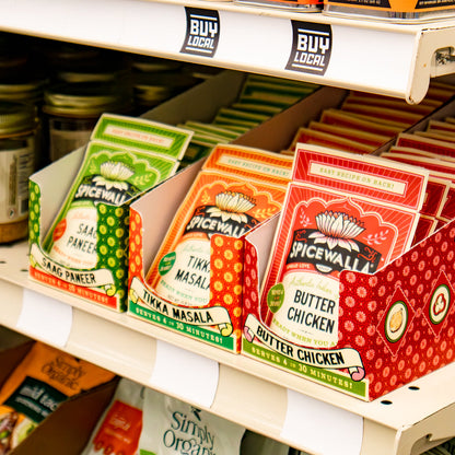 Spicewall seasoning and spice mix products displayed on store shelves, including Spicewalla seasoning tins on the top shelf and Saag Paneer, Tikka Masala, Butter Chicken pouches in front.