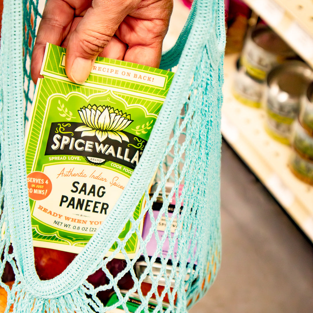 A hand holding a box of Spicewalla Saag Paneer inside a light blue mesh grocery bag, with a blurred supermarket shelf in the background.
