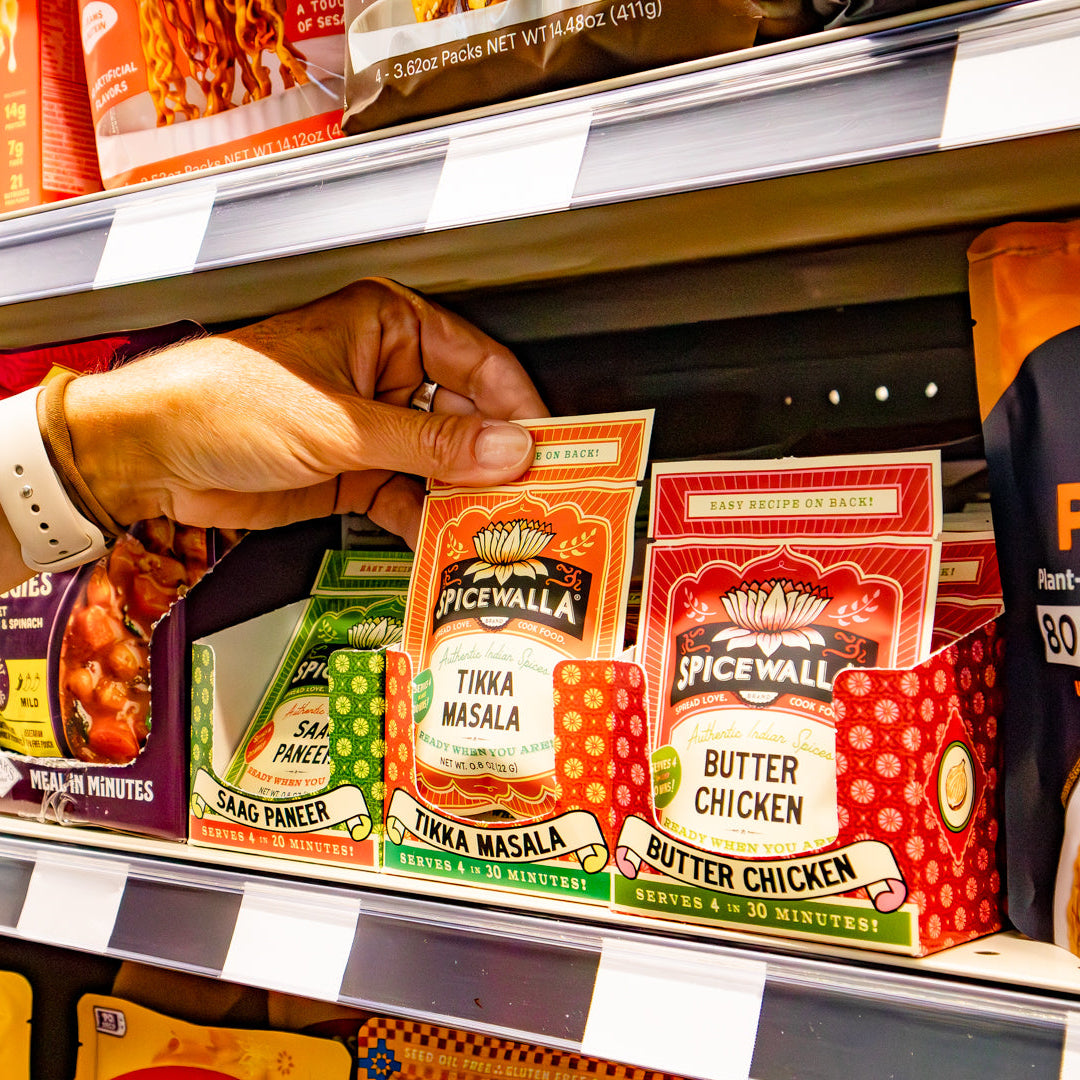 A person's hand reaches for a Spicewall Tikka Masala seasoning packet from a red and white display box on a grocery store shelf.