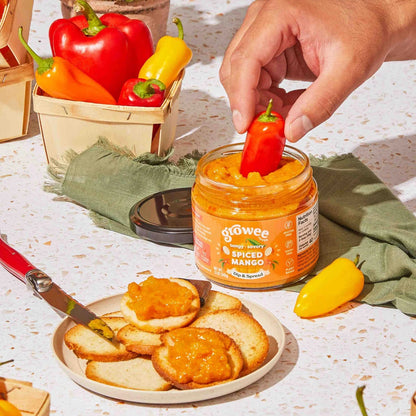 Person dipping a red pepper into a jar of sauce on a table with bread and peppers.