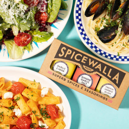 A box of Spicewalla Super Spices & Seasonings sits on a bright turquoise background between three prepared food dishes: a green salad, pasta with mussels, and penne with tomatoes and cheese.