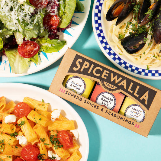 A box of Spicewalla Super Spices & Seasonings sits on a bright turquoise background between three prepared food dishes: a green salad, pasta with mussels, and penne with tomatoes and cheese.