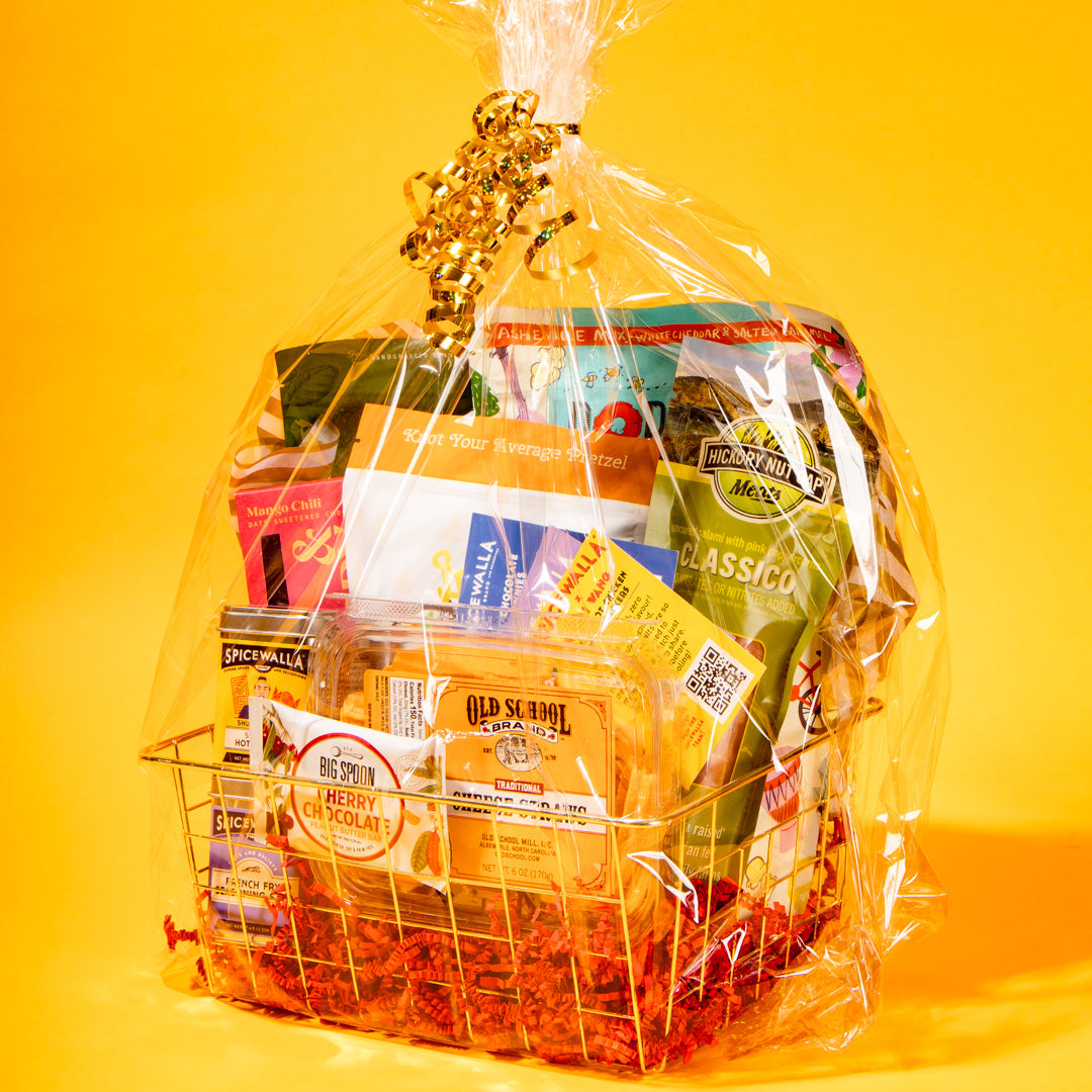 A wire gift basket filled with assorted packaged snacks, crackers, and candy, wrapped in clear cellophane and tied with a gold bow, set against a bright yellow background.
