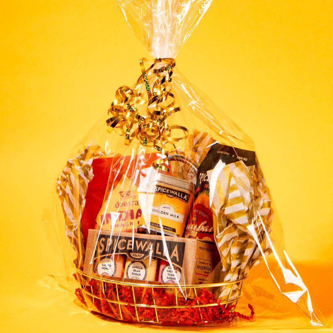 A gift basket wrapped in clear cellophane with a gold ribbon, filled with packaged snacks and food items, set against a solid yellow background.