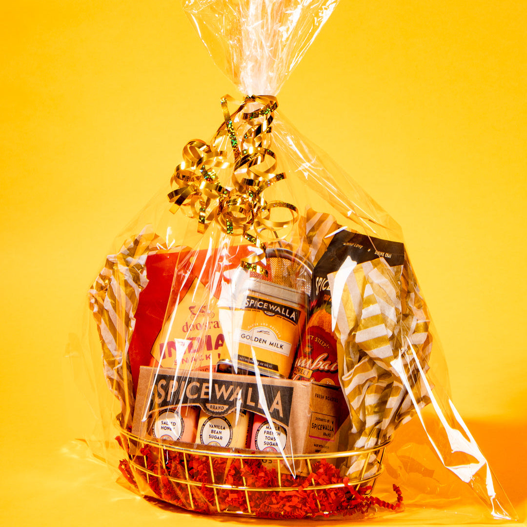 A gift basket wrapped in clear cellophane with a gold ribbon, filled with packaged snacks and food items, set against a solid yellow background.