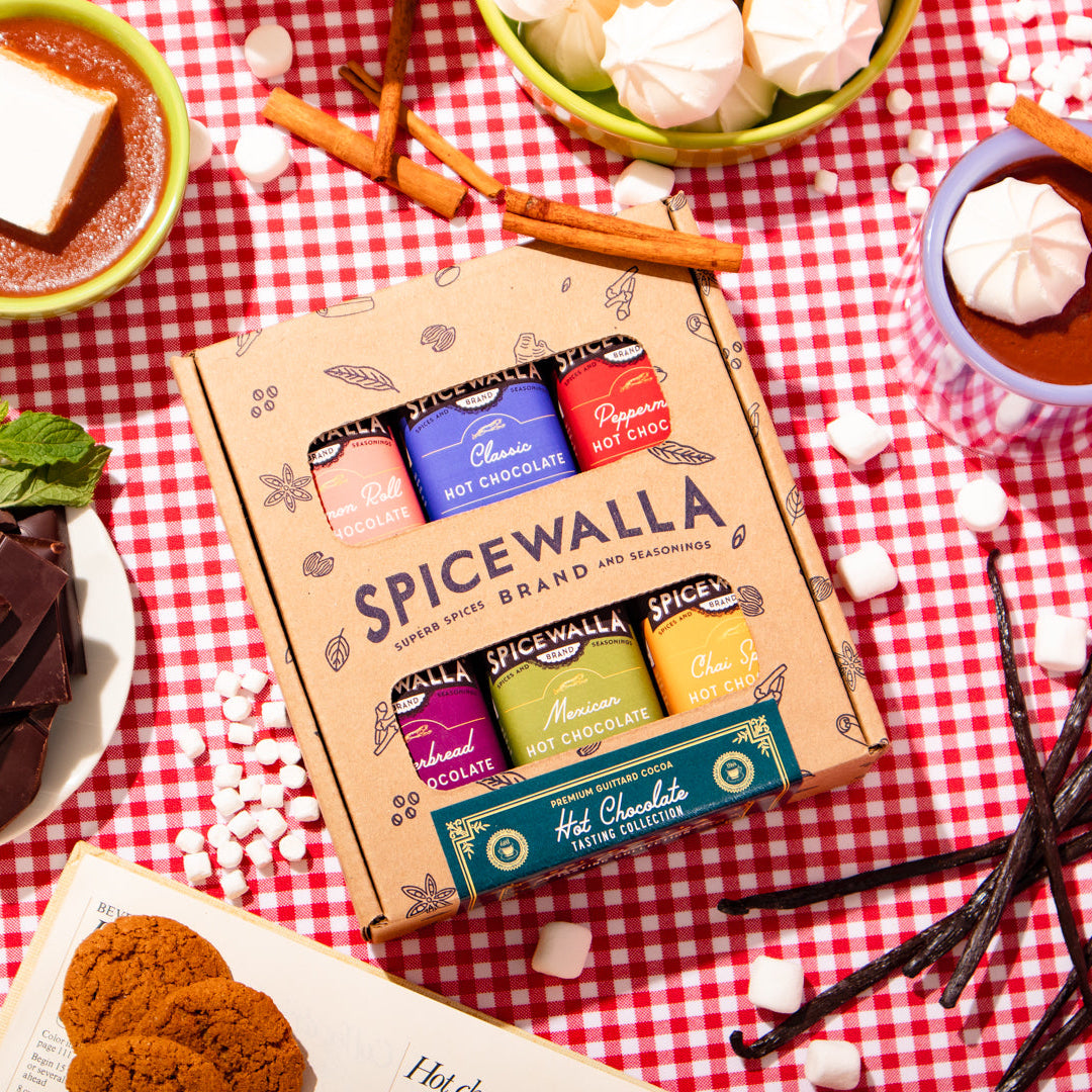 A box of Spicewalla Hot Chocolate Mixtapes coffee-flavored chocolate capsules sits on a red-and-white checkered tablecloth, surrounded by hot cocoa drinks, marshmallows, chocolate chunks, cinnamon sticks, and vanilla beans.