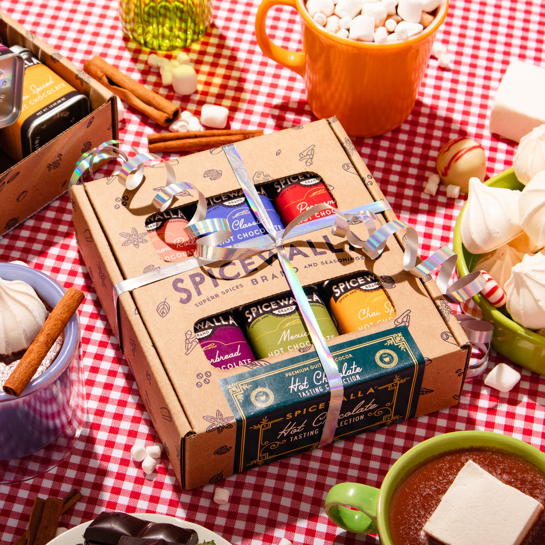A boxed assortment of hot chocolate mix products tied with a silver ribbon, displayed on a red-and-white checkered tablecloth with mugs of hot chocolate, marshmallows, cinnamon sticks, and other holiday-themed food props.