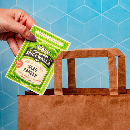 A hand with purple nail polish holds a green and white packet of Spicewalla Saag Paneer above a brown paper shopping bag against a blue geometric background.