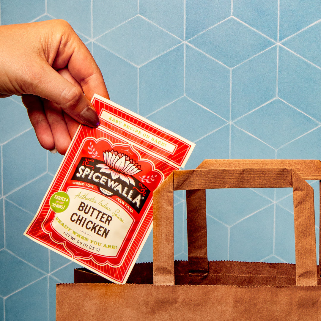 A hand holding a packet of Spicewalla Butter Chicken seasoning above a brown paper shopping bag against a blue geometric tiled background.
