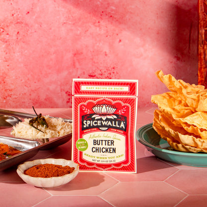 A packet of Spicewalla Butter Chicken seasoning mix stands on a pink surface, surrounded by prepared food including a tray of chicken, rice, a small bowl of red spice, and a plate of golden crispy crackers against a textured pink background.
