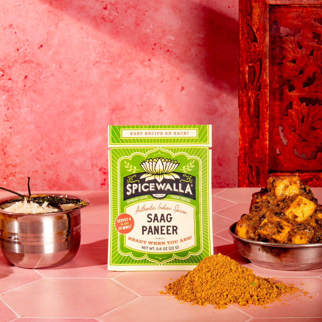 A package of Spicewalla Saag Paneer spice mix is displayed on a pink tiled surface, flanked by a metal bowl of white rice, a pile of yellow-orange ground spice, and a bowl of cooked paneer cubes, all set against a textured pink wall and a carved red wooden panel.