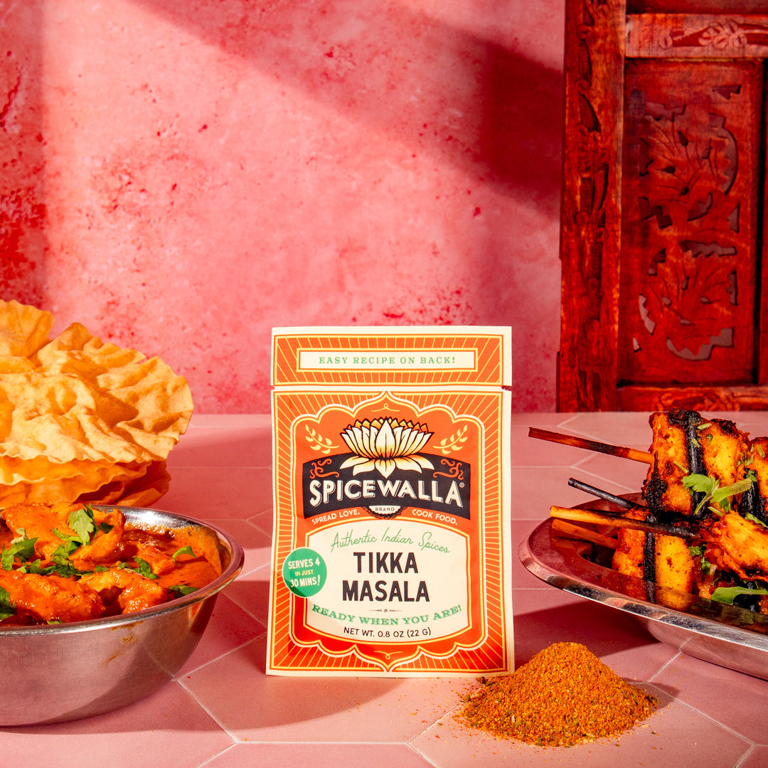A box of Spicewalla Tikka Masala seasoning stands on a pink table, surrounded by cooked chicken skewers, a bowl of curry, and a pile of orange-red spice powder. The background features a textured pink wall and a carved dark red wooden cabinet.