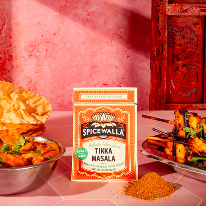 A box of Spicewalla Tikka Masala seasoning stands on a pink table, surrounded by cooked chicken skewers, a bowl of curry, and a pile of orange-red spice powder. The background features a textured pink wall and a carved dark red wooden cabinet.