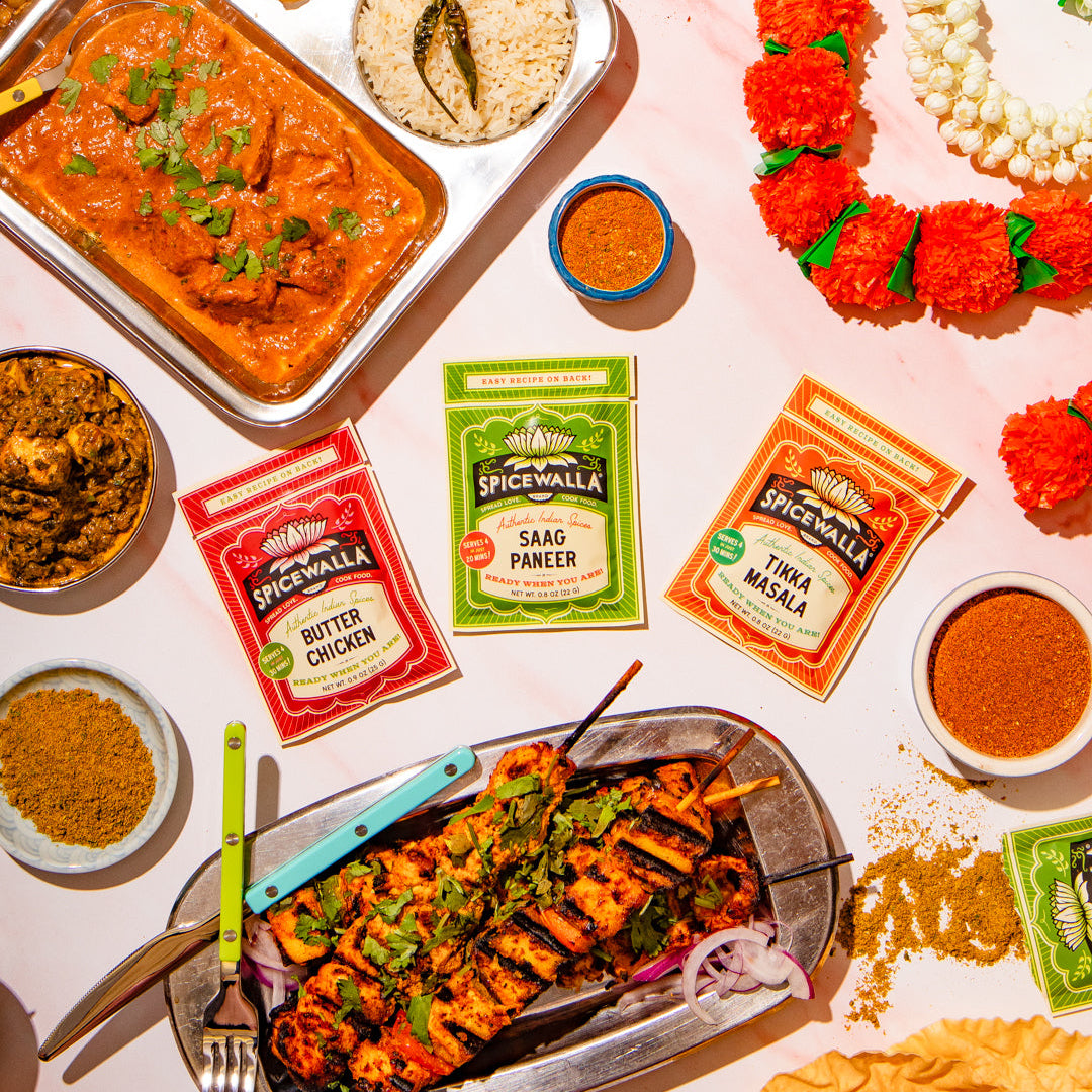 An overhead flat-lay of an Indian spread with grilled skewers, curry, rice, chutney, and three packets of Spicewall seasoning mixes on a light pink surface.