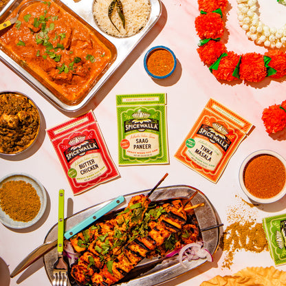 An overhead flat-lay of an Indian spread with grilled skewers, curry, rice, chutney, and three packets of Spicewall seasoning mixes on a light pink surface.