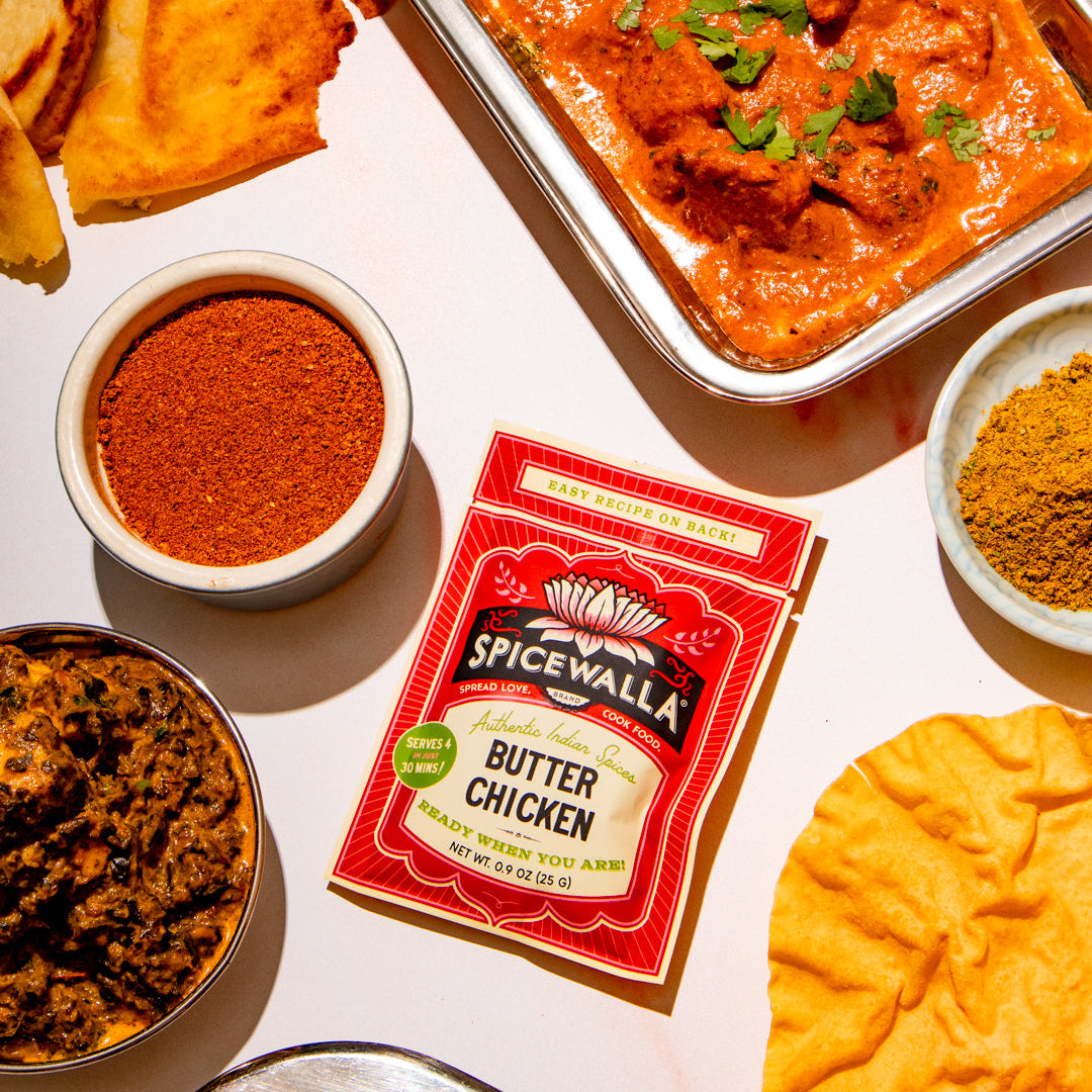 A Spicewalla Butter Chicken seasoning packet lies on a white table surrounded by plated food, including a tray of orange-red chicken curry, flatbread, and small bowls of spices.