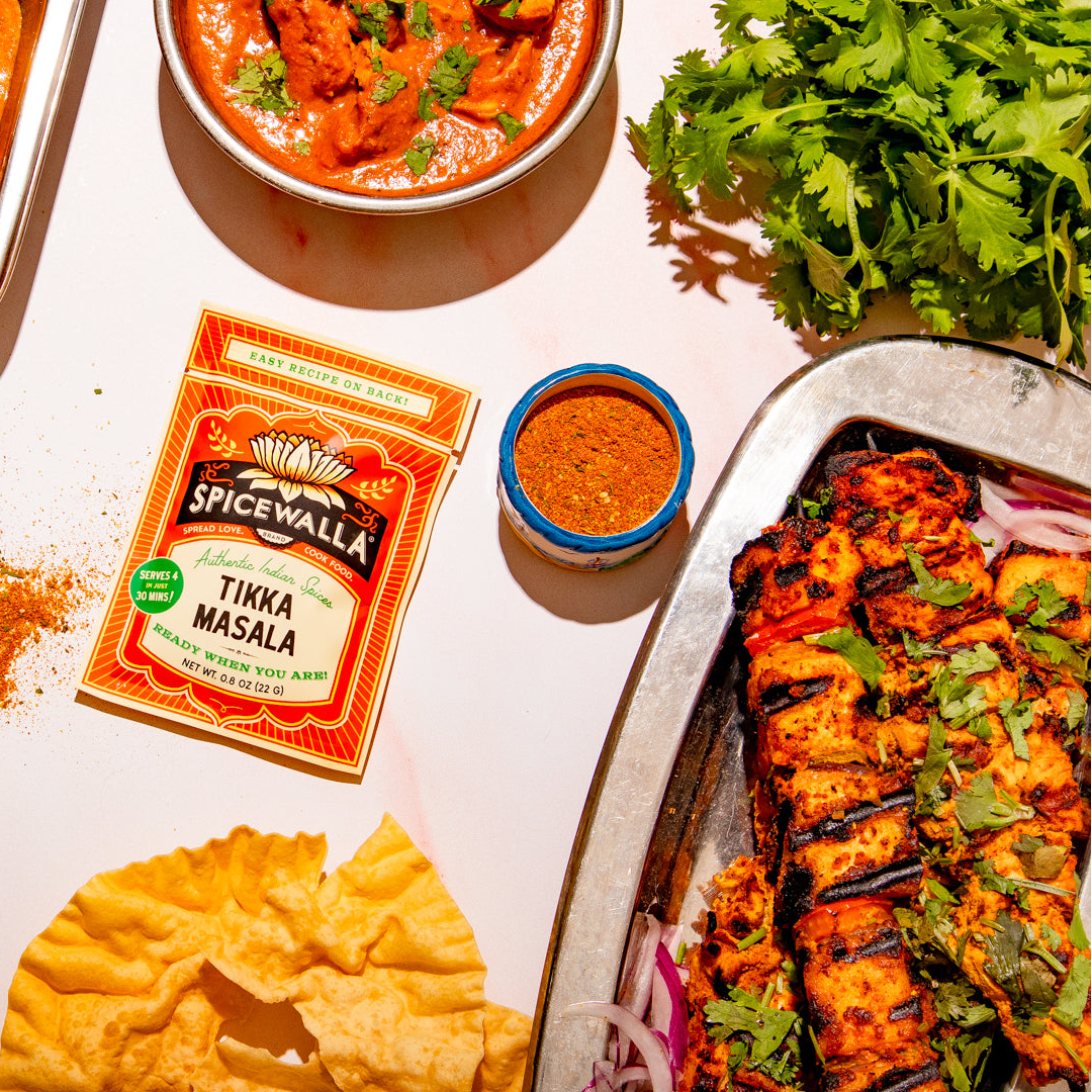 Two packets of Spicewalla Tikka Masala seasoning displayed on a white table alongside prepared food, including a tray of tikka skewers, a bowl of curry, flatbread, and a small bowl of red dipping sauce.