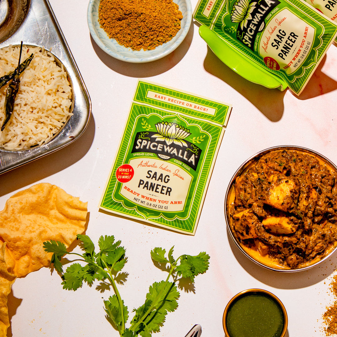 Spicevalla Saag Paneer seasoning mix shown with its green and white packaging, surrounded by prepared food including paneer, flatbread, cilantro, and a small bowl of green chutney on a light surface.