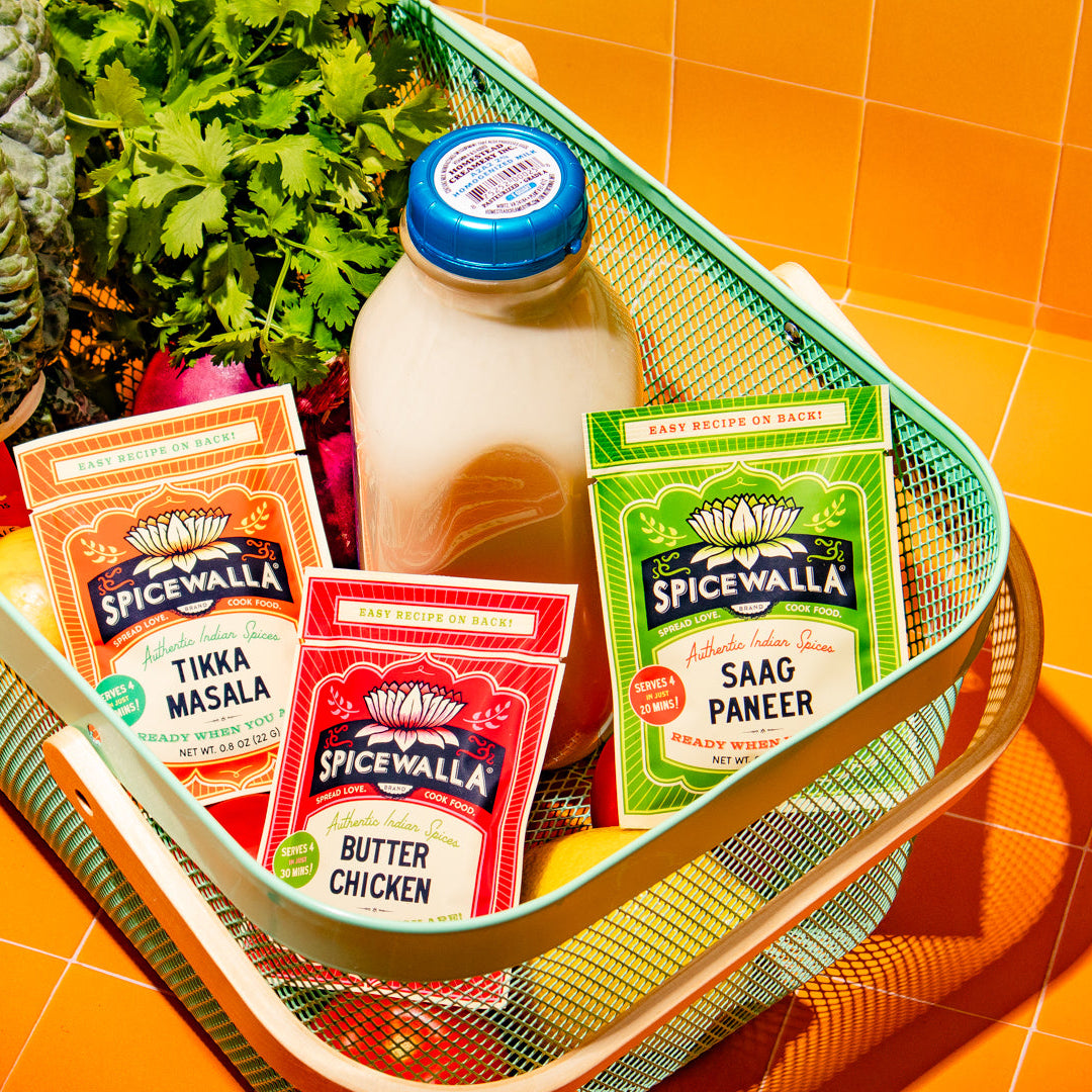 A styled product shot of Spicewalla chicken seasoning packets arranged in a green wire basket with a glass bottle of milk, fresh herbs, and vegetables on an orange tiled background.