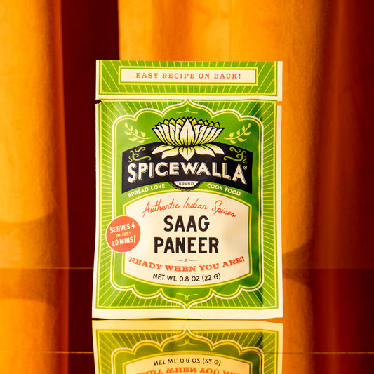 A box of Spicewalla Saag Paneer authentic Indian spices, featuring a green and cream label with a lotus flower illustration, set against a warm orange background with a reflection below.