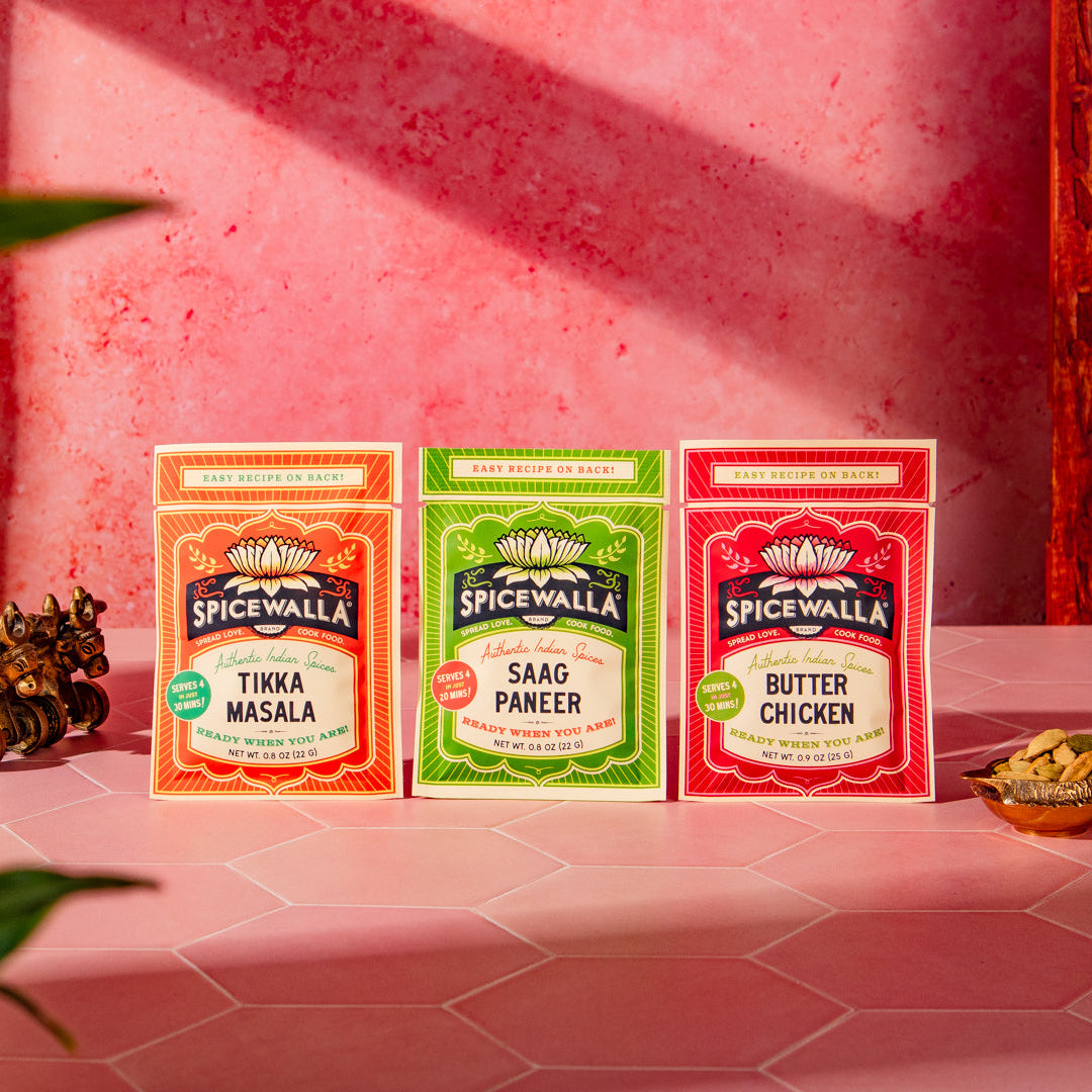 Three SpiceWalla seasoning packets standing upright on a pink surface against a pink wall, with decorative props including a brass cart and leaves in the foreground. The boxes are labeled Tikka Masala, Saag Paneer, and Butter Chicken.