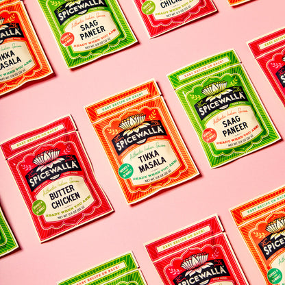 Multiple packets of Spicewall spice mix arranged diagonally on a solid pink background. The packets include Butter Chicken, Sagg Paneer, Tikka Masala, and Tandoori Chicken in red, green, and orange packaging.