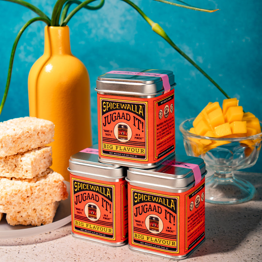 Three tins of 'Spicewell Jugaad It' on a surface with a blue background and decorative elements.
