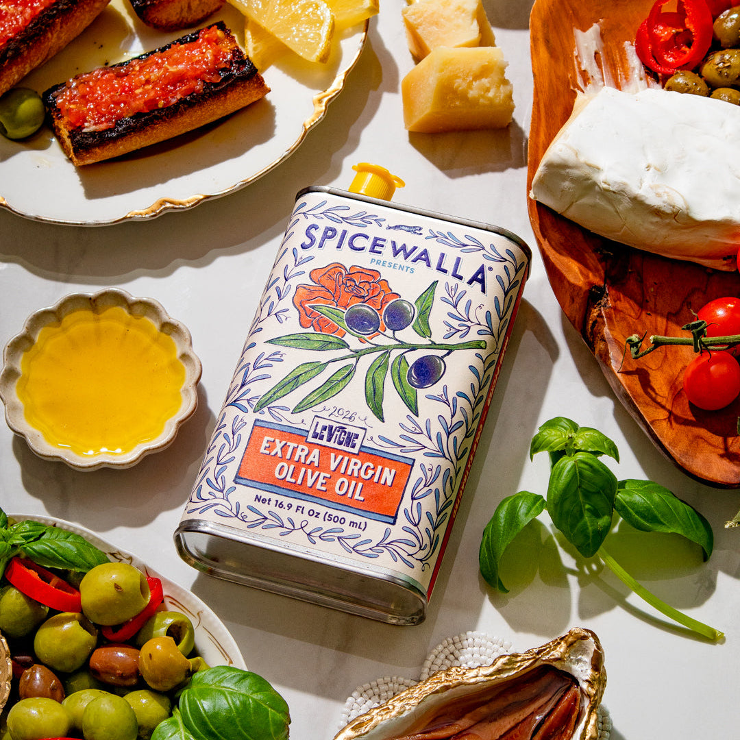 A bottle of Spicewalla extra virgin olive oil is styled in a flat lay with Mediterranean food props including grilled salmon, fresh basil, olives, bread, and cheese on a white surface.
