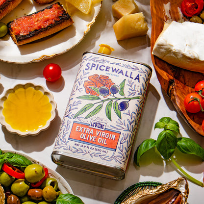 A tin of Spicewalla Extra Virgin Olive Oil is styled in a Mediterranean food setting on a white surface, surrounded by fresh basil, tomatoes, olives, cheese, and grilled bread.