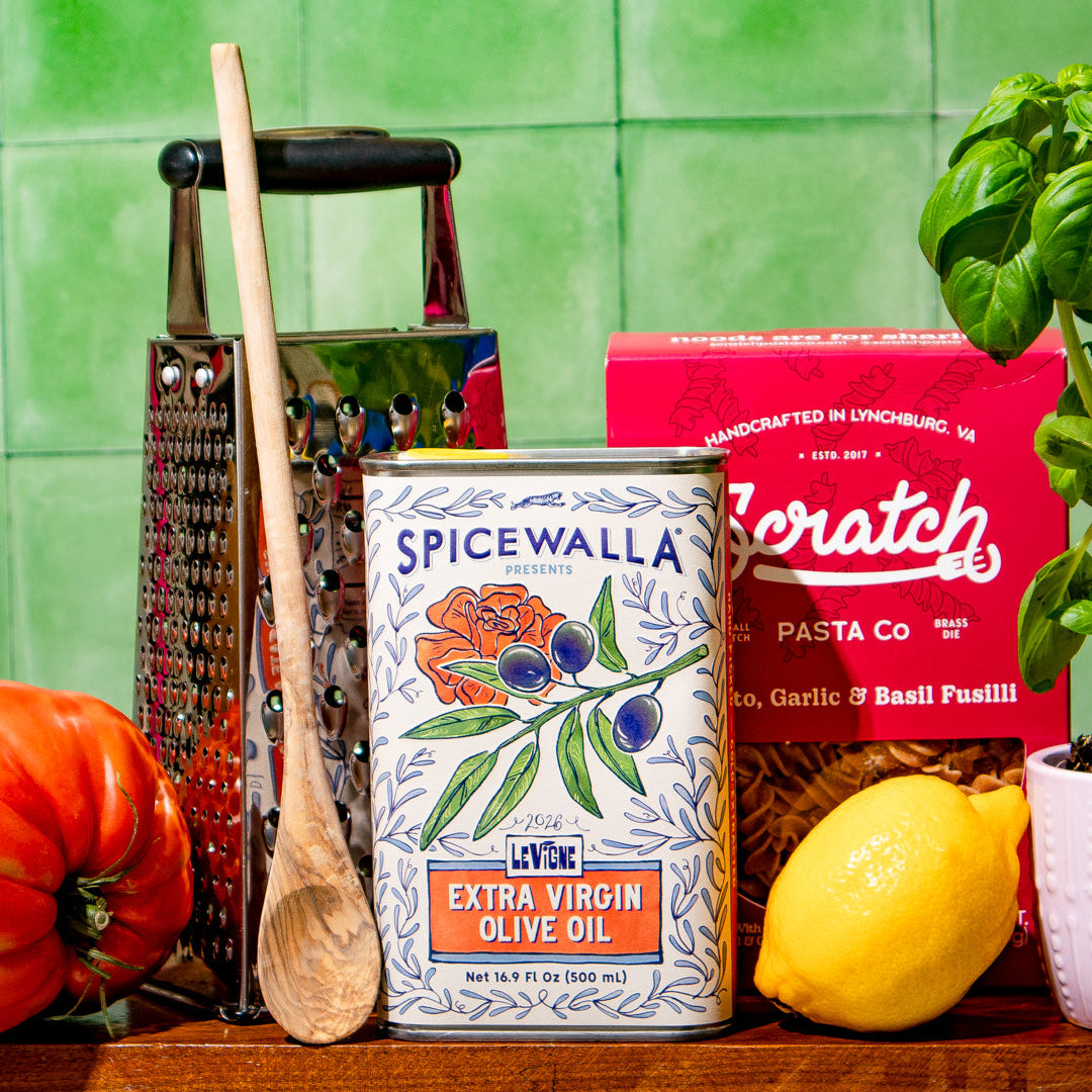 A styled kitchen scene featuring a can of Spicewalla extra virgin olive oil in the center, flanked by a red box of Scratch pasta, a wooden grater, a wooden spoon, a red tomato, a lemon, and a potted basil plant against a green tiled wall.