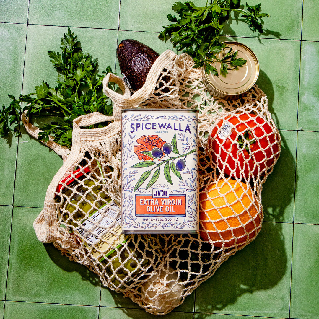A styled flat-lay of a Spicewalla Extra Virgin Olive Oil canister placed on a beige mesh grocery bag, surrounded by fresh ingredients including an avocado, tomatoes, an orange, parsley, and a jar lid, all set on a green tiled surface.