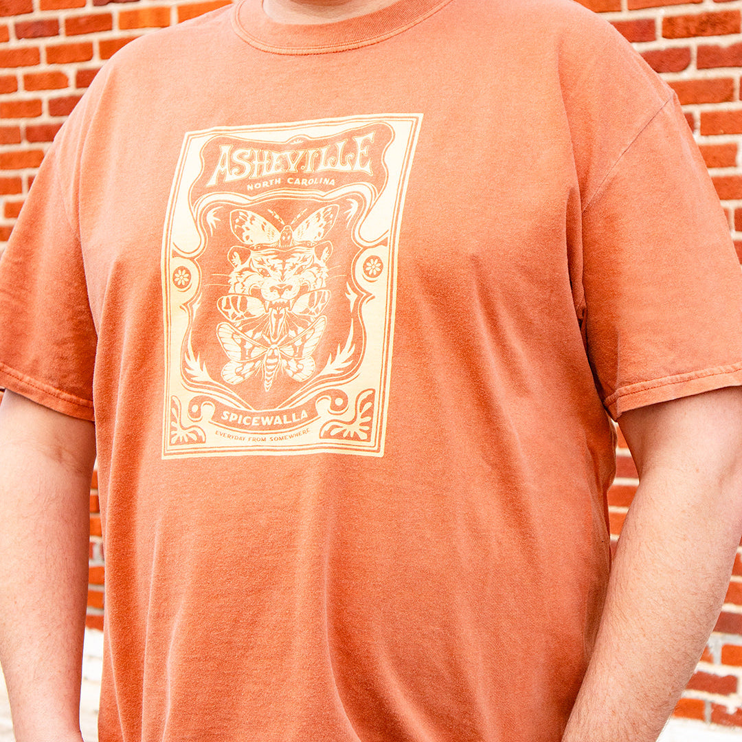 Person wearing an orange t-shirt with a graphic design in front of a brick wall.