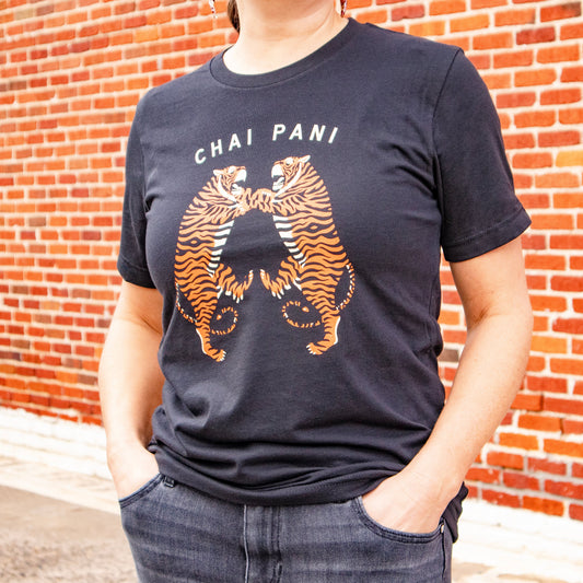 Person wearing a black t-shirt with a tiger graphic and 'Chai Pani' text against a brick wall.