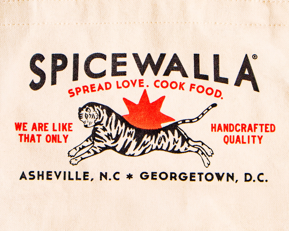 Close-up of a beige canvas tote bag printed with the Spicewalla brand logo, featuring black text, red slogan text, and a black-and-white tiger graphic on a cream fabric background.