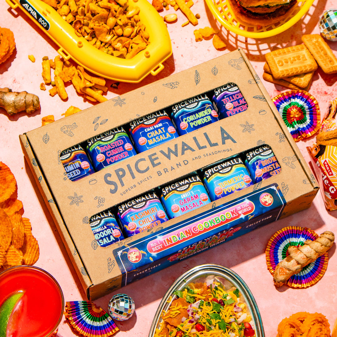A Spicewalla gift box filled with assorted canned sodas and drinks, surrounded by colorful food props like a burger, tortilla chips, a drink with lime, and a loaded taco dish on a pink surface.