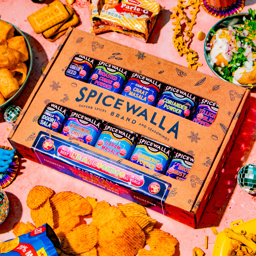 A Spicewalla snack assortment box displayed on a pink surface surrounded by various snacks, chips, and drinks.