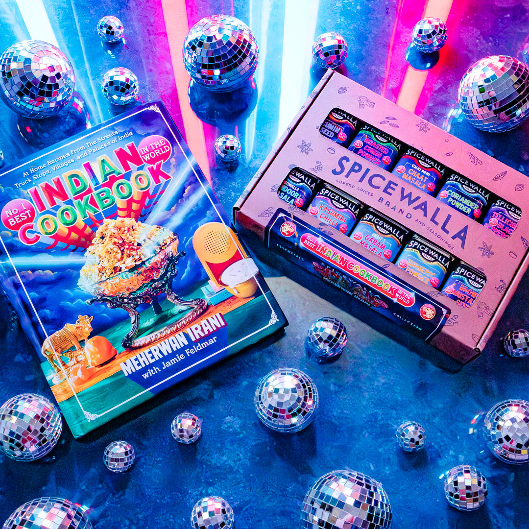 A boxed set of colorful spice blends and a cookbook titled 'Indian Cookbook' arranged on a glossy blue surface with many mirrored disco balls. The box reads 'Spicewalla' and shows multiple spice packets inside; the book cover features bright food photography and the text 'Merewah Iranian.
