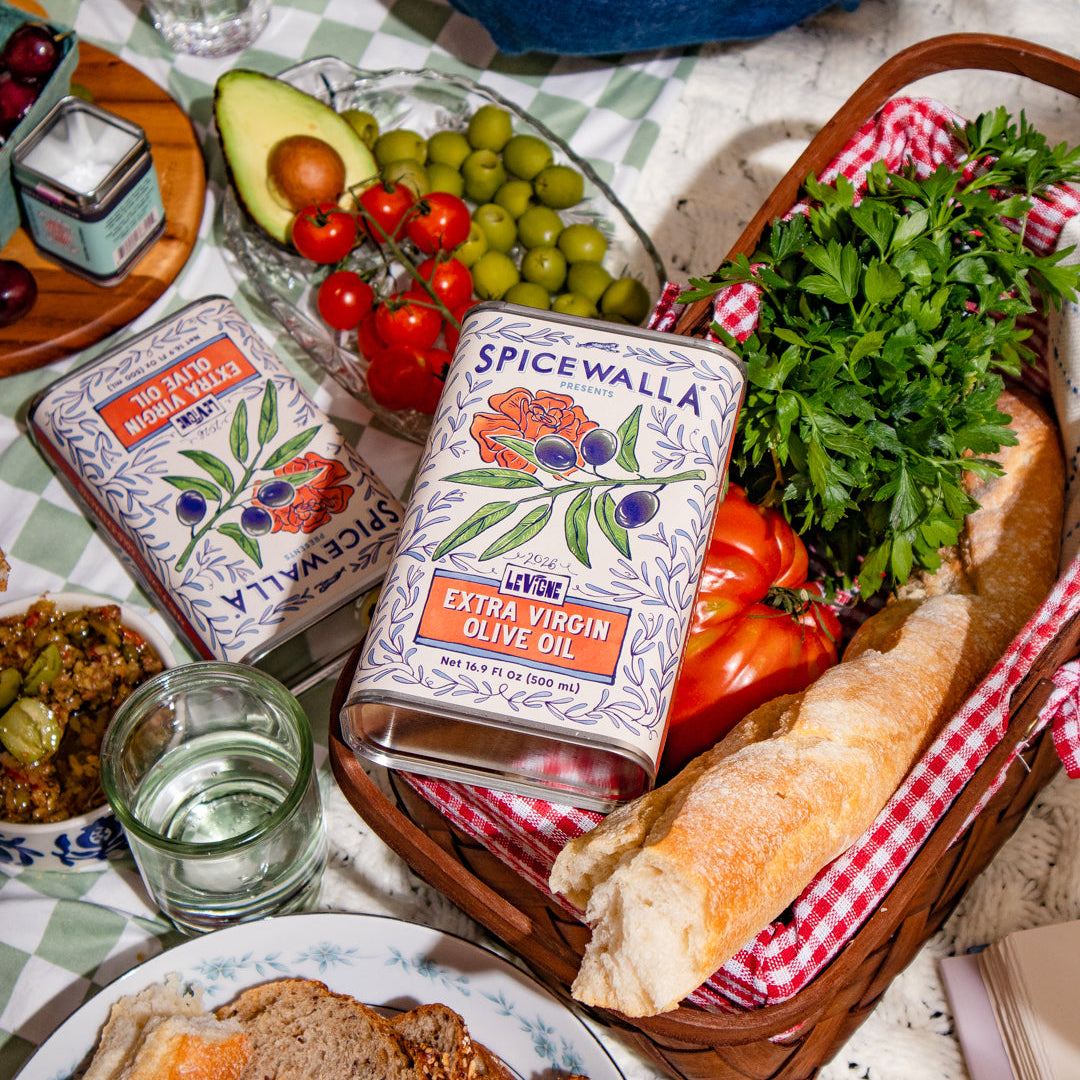 Two Spicewalla Extra Virgin Olive Oil tins displayed on a rustic tablecloth with food props including sliced bread, fresh parsley, tomatoes, olives, and a baguette in a basket.