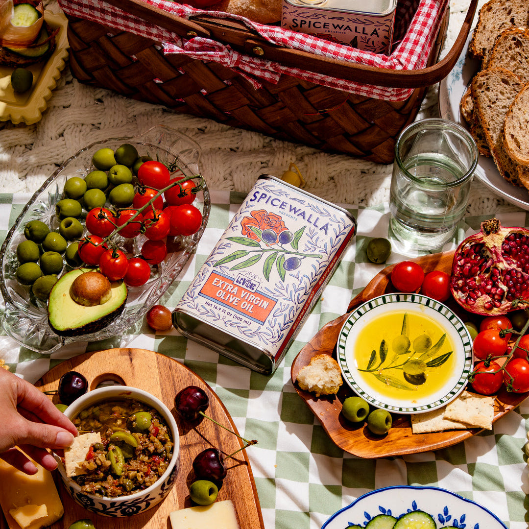 A styled tabletop spread featuring a woven basket with bread and a can of Spiced Walls Mediterranean Sausage, alongside olives, tomatoes, cheese, cured meats, a bowl of olive oil, sliced cucumbers, and a pomegranate on a checkered tablecloth.